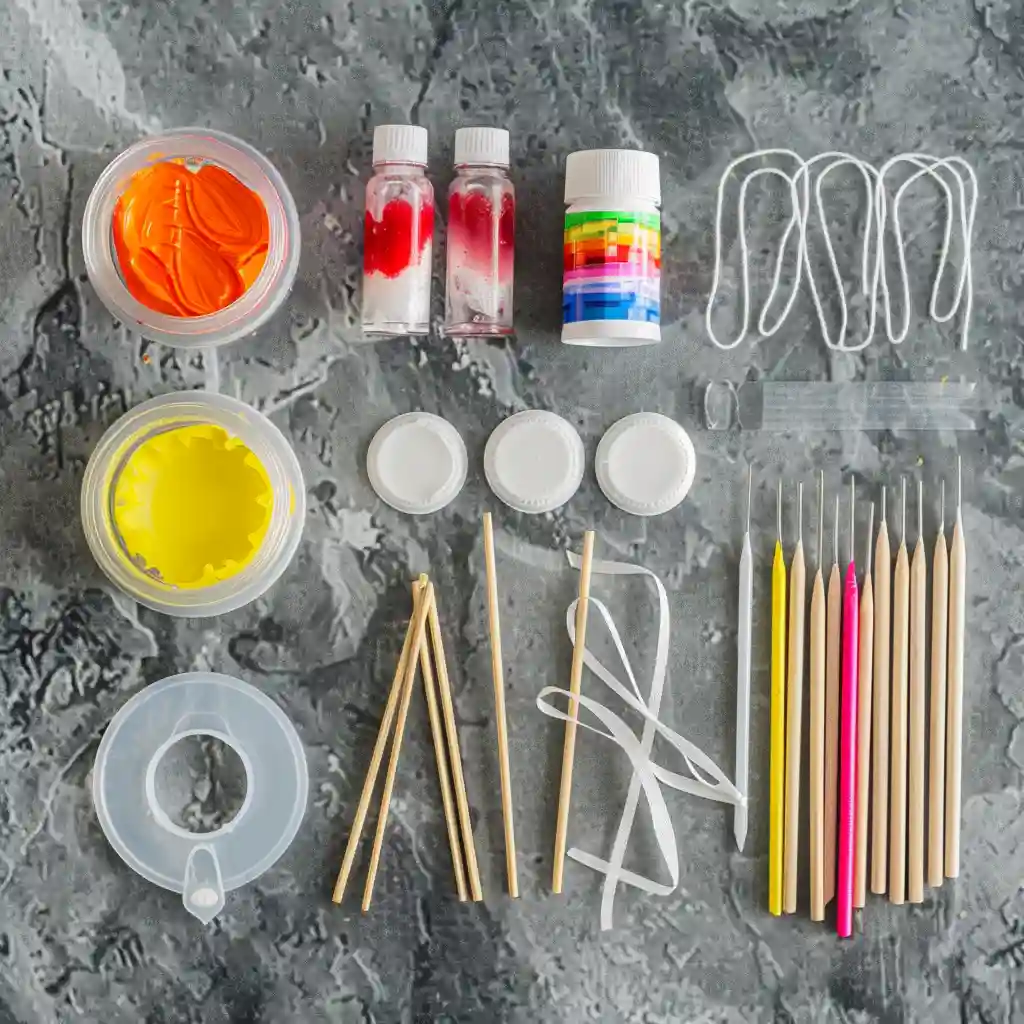 DIY suncatcher craft kit with paints, sticks, ribbons, and sculpting tools arranged on a textured gray surface.